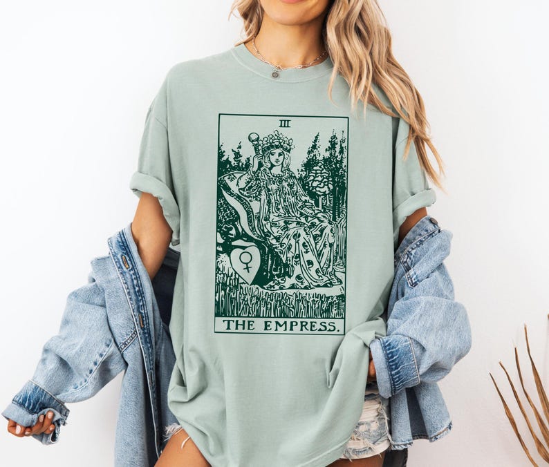 May include: A light green t-shirt with a graphic of the Empress tarot card. The card features a woman sitting on a throne with a crown on her head. The text "THE EMPRESS" is printed below the card.