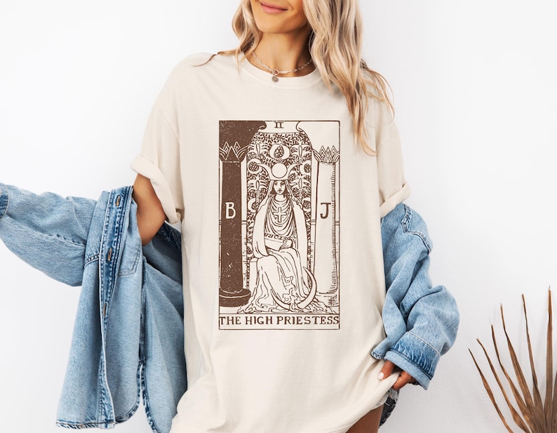 May include: A beige t-shirt with a brown graphic of the High Priestess tarot card. The card features a woman sitting on a throne with a crown and a crescent moon above her head. The text "The High Priestess" is printed below the card.