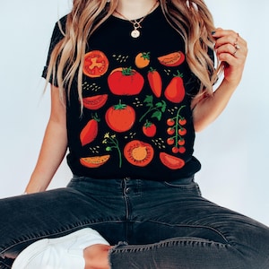 May include: A black t-shirt featuring a red, orange, and green tomato print. The shirt depicts various sizes and shapes of tomatoes, including whole tomatoes, tomato slices, and cherry tomatoes.