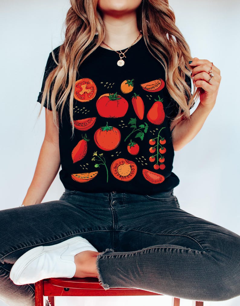 May include: A black t-shirt featuring a red, orange, and green tomato print. The shirt depicts various sizes and shapes of tomatoes, including whole tomatoes, tomato slices, and cherry tomatoes.