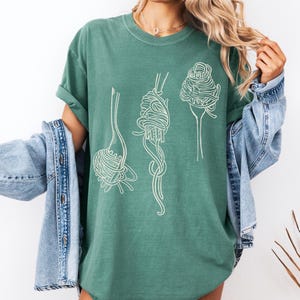 K&ouml;nnte beinhalten: Ein gr&uuml;nes T-Shirt mit einer wei&szlig;en Grafik von drei Gabeln mit Spaghetti darauf.