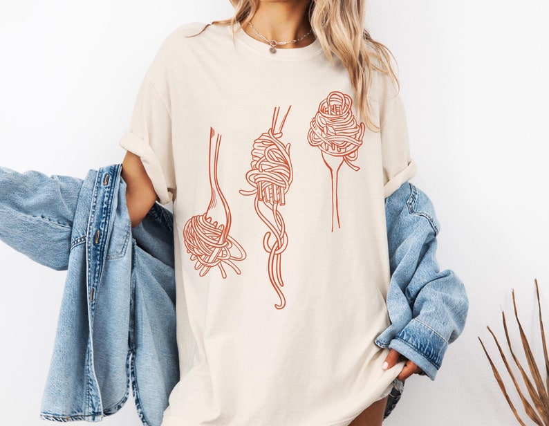 K&ouml;nnte beinhalten: Ein wei&szlig;es T-Shirt mit einem Motiv aus drei Spaghetti-Nudeln auf Gabeln. Die Nudeln sind in rot-orange gezeichnet.