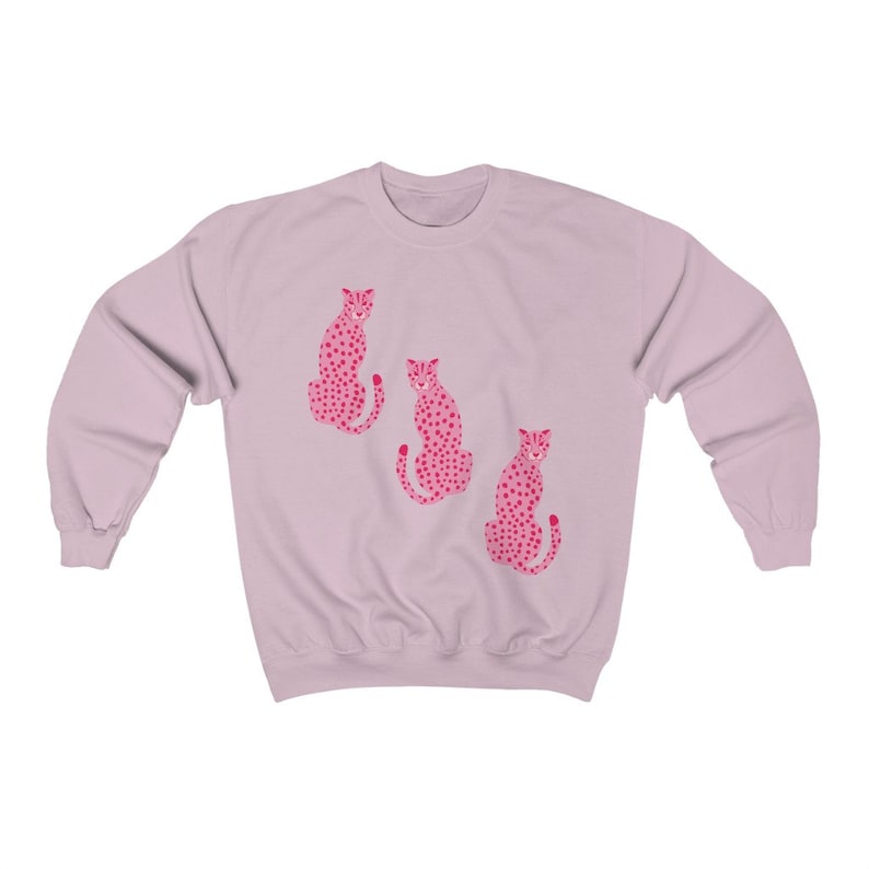 May include: A light pink sweatshirt with a graphic print of three pink leopards with black spots. The leopards are sitting in a row, facing forward.