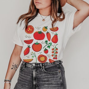 May include: White t-shirt with a colourful tomato print. The print features red, orange, and green tomatoes in various shapes and sizes.