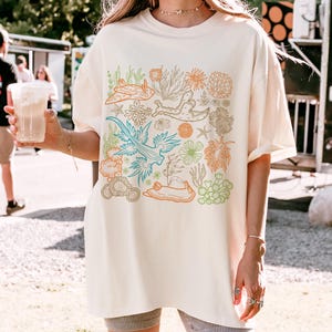 Pu&ograve; includere: Una t-shirt color crema con una stampa grafica di lumache di mare colorate e coralli. La grafica &egrave; centrata sulla t-shirt ed &egrave; circondata da un bordo bianco.