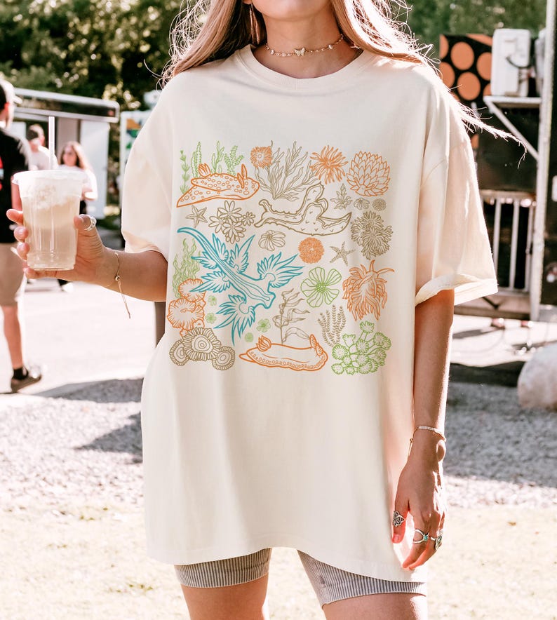 Pu&ograve; includere: Una t-shirt color crema con una stampa grafica di lumache di mare colorate e coralli. La grafica &egrave; centrata sulla t-shirt ed &egrave; circondata da un bordo bianco.
