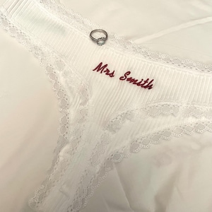 Puede incluir: Tanga blanco con textura acanalada y delicado ribete de encaje. El texto "Mrs. Smith" está bordado en hilo burdeos. Un anillo plateado con una piedra transparente descansa sobre la ropa interior, sugiriendo un tema nupcial.