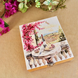 Puede incluir: Caja de madera con una pintura de una mesa con vino, queso e higos en un balcón. La obra de arte presenta una botella de vino tinto, copas y flores rosas en primer plano.