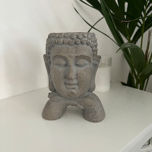 May include: A grey concrete planter shaped like a meditating Buddha head. The planter has a small opening at the top for planting.