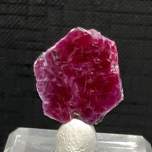 May include: A deep red ruby crystal with a hexagonal shape. The crystal is mounted on a white stand.