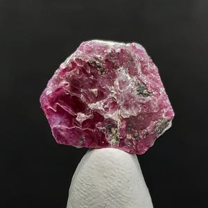 Può includere: Una gemma di rubino grezza, non tagliata, con una tonalità rosa intenso, che mostra una struttura cristallina e imperfezioni naturali. Il rubino è montato su una base bianca e arrotondata su sfondo nero. La superficie della pietra riflette la luce.