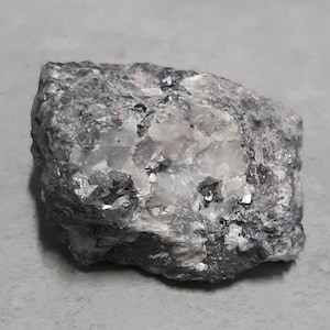 Può includere: Primo piano di una roccia grigia ruvida con cristalli incrostati. I cristalli sono un mix di bianco e nero, alcuni con un bagliore metallico.