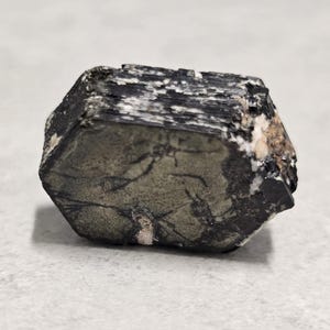 May include: A dark, multi-faceted mineral specimen with a mix of black, gray, and white colors. The stone has a rough, textured surface and a geometric shape. The mineral appears to be a natural, unpolished stone.