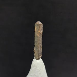 May include: A natural, brown crystal with a pointed tip. The crystal is a light brown color with some darker brown and orange hues. It is standing upright on a white base against a black background.