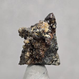May include: A close-up of a mineral specimen with a rough, textured surface. The mineral displays a mix of dark gray, brown, and gold colors, with a crystalline structure. The specimen is mounted on a white base.