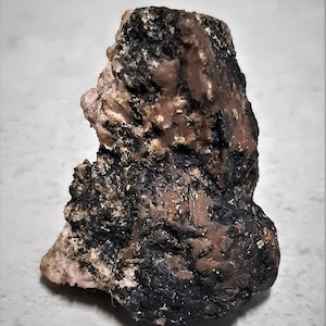 May include: A rough, irregularly shaped rock with a mix of colors. The rock has areas of black, brown, and pinkish-tan hues. The texture appears coarse and uneven, with a slightly glossy sheen.