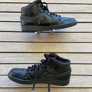 Puede incluir: Un par de zapatillas de deporte de caña alta de cuero negro con suelas blancas. Las zapatillas están colgadas de ganchos en una pared de madera.