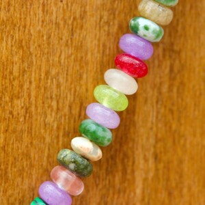 May include: A close-up of a colorful beaded necklace. The beads are oval-shaped and come in a variety of colors, including green, purple, red, and white. The necklace is displayed against a wooden background.