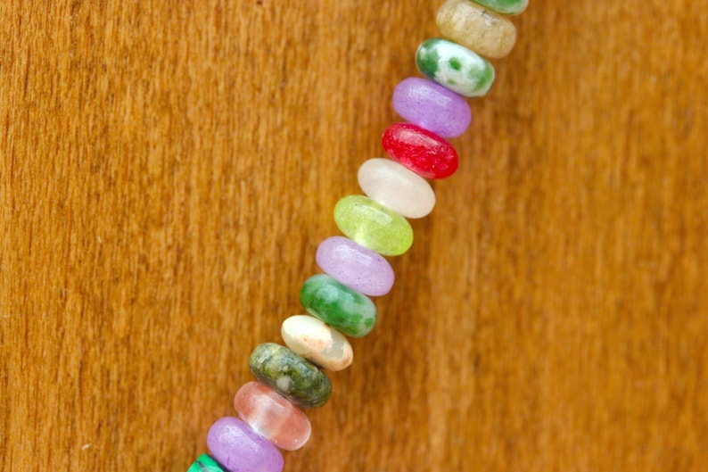 May include: A close-up of a colorful beaded necklace. The beads are oval-shaped and come in a variety of colors, including green, purple, red, and white. The necklace is displayed against a wooden background.