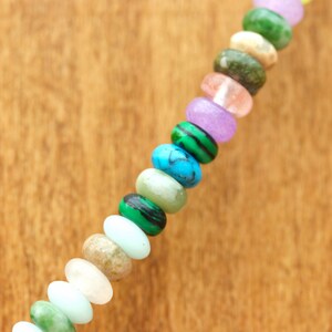 May include: A close-up of a necklace featuring a variety of colorful gemstone beads. The beads are oval-shaped and include shades of green, blue, pink, purple, and white. The necklace is set against a warm, wooden background.