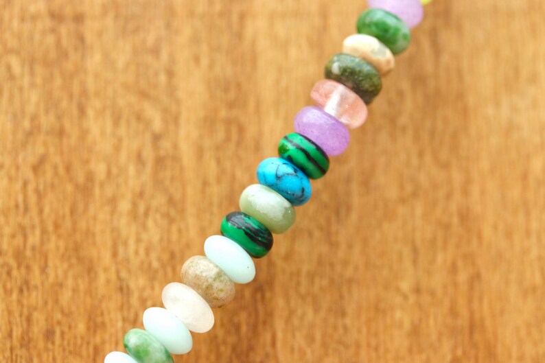 May include: A close-up of a necklace featuring a variety of colorful gemstone beads. The beads are oval-shaped and include shades of green, blue, pink, purple, and white. The necklace is set against a warm, wooden background.