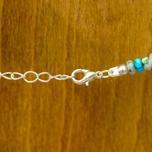 May include: Close-up of a necklace with a silver chain and clasp. The necklace features a strand of colorful beads, including turquoise, green, white, and red. The background is a warm, wooden surface.