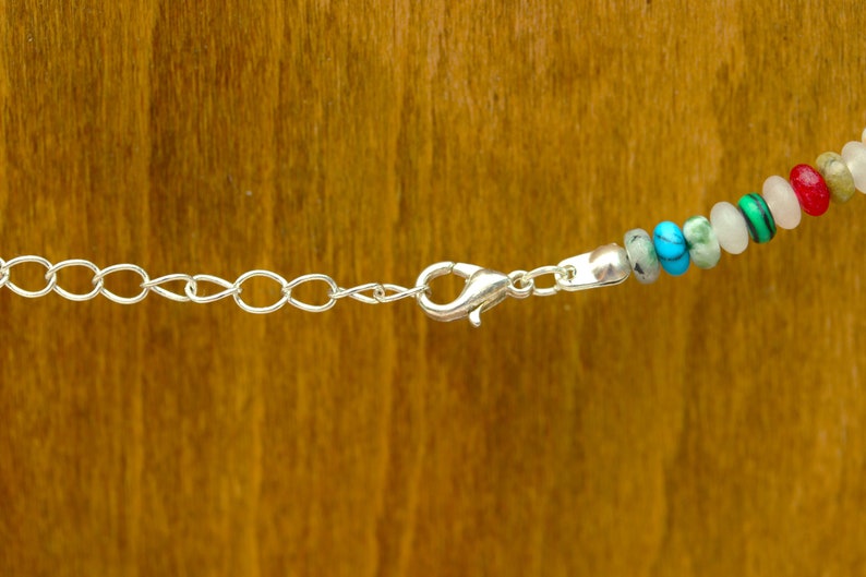 May include: Close-up of a necklace with a silver chain and clasp. The necklace features a strand of colorful beads, including turquoise, green, white, and red. The background is a warm, wooden surface.