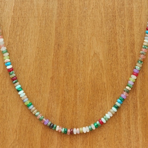 May include: A colorful beaded necklace with a variety of small, round gemstones in shades of red, green, blue, pink, and white. The necklace is displayed on a wooden stand, showcasing the vibrant colors and unique design of the jewelry.