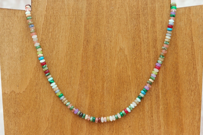May include: A colorful beaded necklace with a variety of small, round gemstones in shades of red, green, blue, pink, and white. The necklace is displayed on a wooden stand, showcasing the vibrant colors and unique design of the jewelry.
