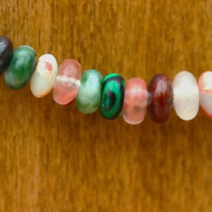 May include: Close-up of a beaded necklace with a variety of colorful, oval-shaped beads. The beads are in shades of purple, green, brown, white, and pink. The necklace is set against a warm, wooden background.