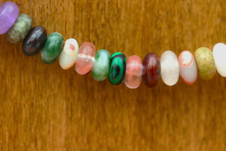 May include: Close-up of a beaded necklace with a variety of colorful, oval-shaped beads. The beads are in shades of purple, green, brown, white, and pink. The necklace is set against a warm, wooden background.
