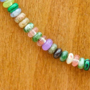 May include: A close-up of a necklace with colorful, oval-shaped beads. The beads are a mix of colors including green, pink, purple, and brown. The necklace is set against a wooden background.