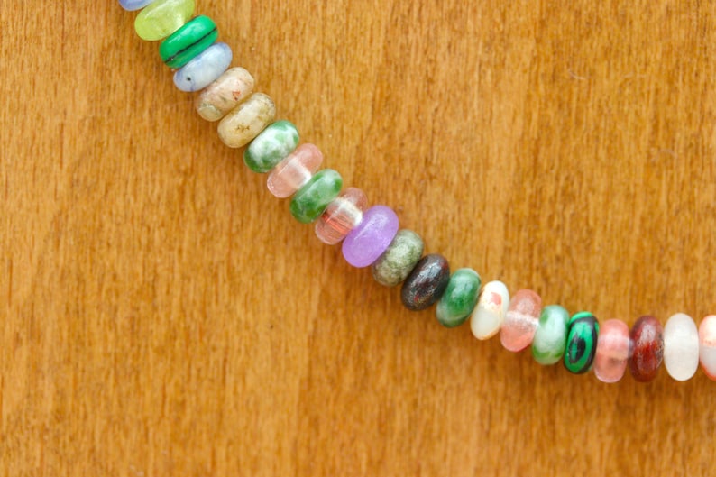 May include: A close-up of a necklace with colorful, oval-shaped beads. The beads are a mix of colors including green, pink, purple, and brown. The necklace is set against a wooden background.