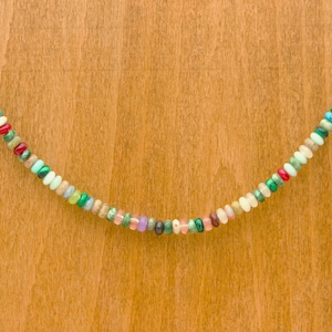 May include: A colorful beaded necklace with a variety of small, round beads in shades of red, green, blue, pink, and white. The necklace is displayed against a light brown wooden background.