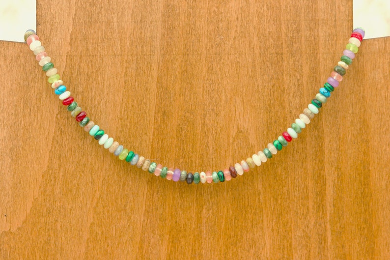 May include: A colorful beaded necklace with a variety of small, round beads in shades of red, green, blue, pink, and white. The necklace is displayed against a light brown wooden background.