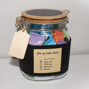 May include: A glass jar with a black lid and a label that says "Jar of date ideas". The jar is filled with colourful paper squares. The label also has a list of categories for the date ideas: Fun & Free, Days out, Relaxing, Adventure, and Special.