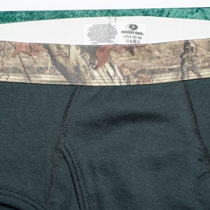 May include: Close-up of black boxer briefs with a camouflage waistband. The waistband features a brown, green, and tan pattern. The tag reads "Mossy Oak" and "L/G (36-38)".