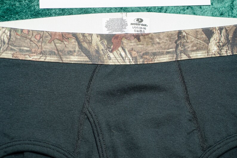 May include: Close-up of black boxer briefs with a camouflage waistband. The waistband features a brown, green, and tan pattern. The tag reads "Mossy Oak" and "L/G (36-38)".