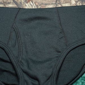 May include: Black briefs with a camouflage waistband. The waistband features a brown and green camouflage pattern. The tag reads "Mossy Oak" and "L/G/G (36-38)".