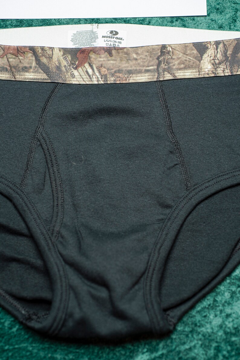 May include: Black briefs with a camouflage waistband. The waistband features a brown and green camouflage pattern. The tag reads "Mossy Oak" and "L/G/G (36-38)".