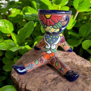 May include: A colorful ceramic planter with a unique, whimsical design. The planter features a wide, bowl-shaped top with a vibrant floral pattern in red, yellow, and blue. It rests on three brown legs with blue accents, creating a playful, artistic look.