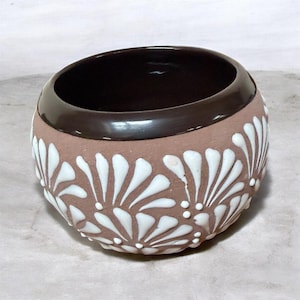 May include: A small, round ceramic bowl with a brown interior and rim. The exterior is a light brown with a white, stylized floral pattern. The bowl has a smooth, glossy finish.