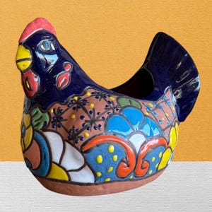 May include: A ceramic chicken-shaped planter featuring a glossy dark blue head and tail with a red comb and yellow beak. The body displays a terracotta base color decorated with vibrant hand-painted floral patterns in blue, yellow, orange, and white.