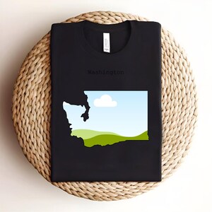 May include: Black t-shirt featuring a graphic of the Washington state outline with a landscape design inside. The design includes a blue sky, white cloud, and green hills. The word "Washington" is printed above the graphic.