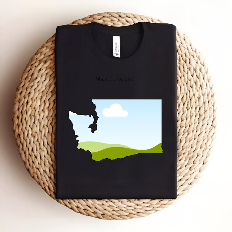 May include: Black t-shirt featuring a graphic of the Washington state outline with a landscape design inside. The design includes a blue sky, white cloud, and green hills. The word "Washington" is printed above the graphic.