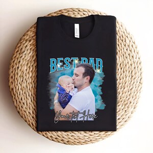 May include: Black t-shirt with a blue and white graphic that says "Best Dad" with a photo of a father and child. The text "Your Text Here" is below the graphic.