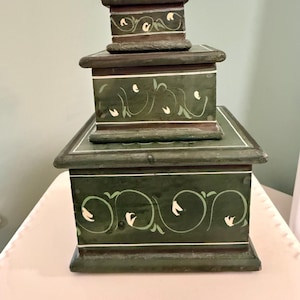 May include: Three-piece nesting box set. The boxes are painted dark green with white floral and vine designs. The boxes are stacked on top of each other, with the smallest box on top.