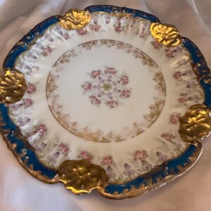 May include: A white porcelain plate with a blue rim and gold accents. The plate features a floral pattern in the center and around the edges. Gold-colored decorative elements are placed around the rim.