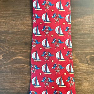 May include: A red necktie featuring a repeating pattern of white sailboats with blue accents, blue dolphins wearing sailor hats, and small yellow stars. The nautical-themed print is evenly distributed across the entire length of the fabric.