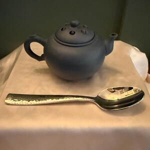 May include: A small, dark blue ceramic teapot with a rounded body, a curved handle, and a lid with several small holes. A silver spoon with a hammered texture lies next to the teapot.